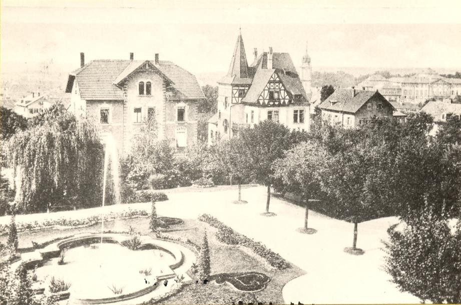 schwarz-weiß Foto des Stadtparks mit Springbrunnen, angelegten Beeten, einer Allee, dahinter drei Villen, noch weiter hinten das Schloss
