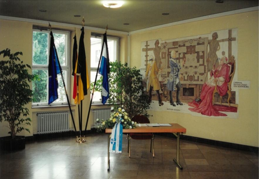 Foto Foyer vor Ratssaal mit vier Fahnen im Hintergrund und Wandgemälde rechts, im Vordergrund Tisch mit Blumenschmuck und Liste.