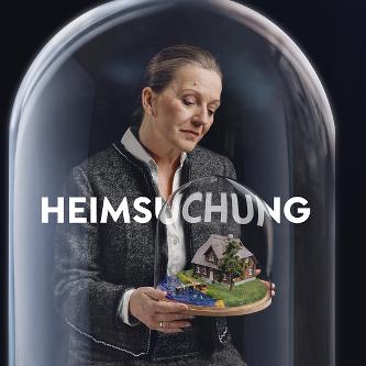 Eine Frau in grauer Strickjacke und Rock sitzt unter einer Glaskuppel. In der Hand hält sie ein Modell von einem Haus am See, ebenfalls unter einer Glaskuppel.
