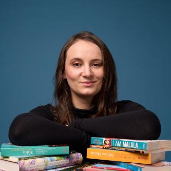 Eine junge Frau sitzt an einem Tisch, die Ellenbogen auf zwei Bücherstapel gestützt. Sie lächelt in die Kamera.