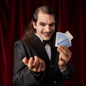Portraitfoto eines Zauberers mit zurück-gegelten, langen Haaren, der einen Kartentrick zeigt. Er sieht sehr unangenehm aus.