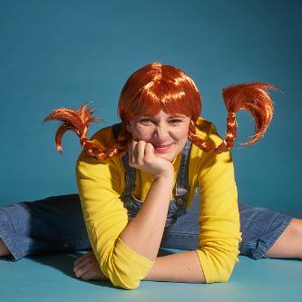 Foto einer jungen Frau als Pippi Langstrumpf verkleidet, mit blauer Latzhose, gelbem Pulli und roten, abstehenden Zöpfen. Sie sitzt im Spagat auf dem Boden, den Kopf auf eine Hand gestützt.