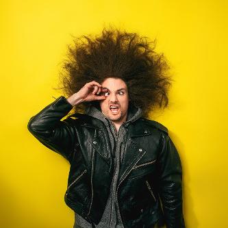 Portraitfoto eines Mannes in Lederjacke vor gelbem Hintergrund. Seine langen Haare stehen in alle Richtungen ab. Er reißt sein rechtes Auge mit zwei Fingern auf und zieht eine Grimasse.