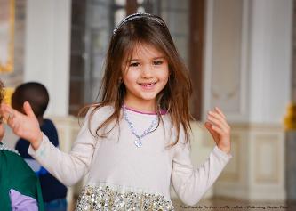 Mädchen mit Diadem und Schmuck lächelt im Schloss Bruchsal in die Kamera. Im Hintergrund sind weitere Kinder beim Ferienspaß zu sehen.
