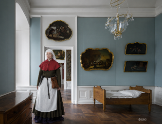 Eine Magd in historischer Kleidung steht in einem barock eingerichteten Dienstbotenzimmer mit Bett, Kommode und Gemälden.